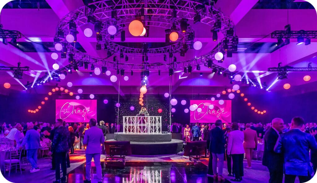 A large crowd fills a purple-lit event hall with hanging round lights, a circular stage centerpiece, and two screens displaying the word “Chase.” People mingle in formal attire under dramatic lighting and a modern ceiling structure.
