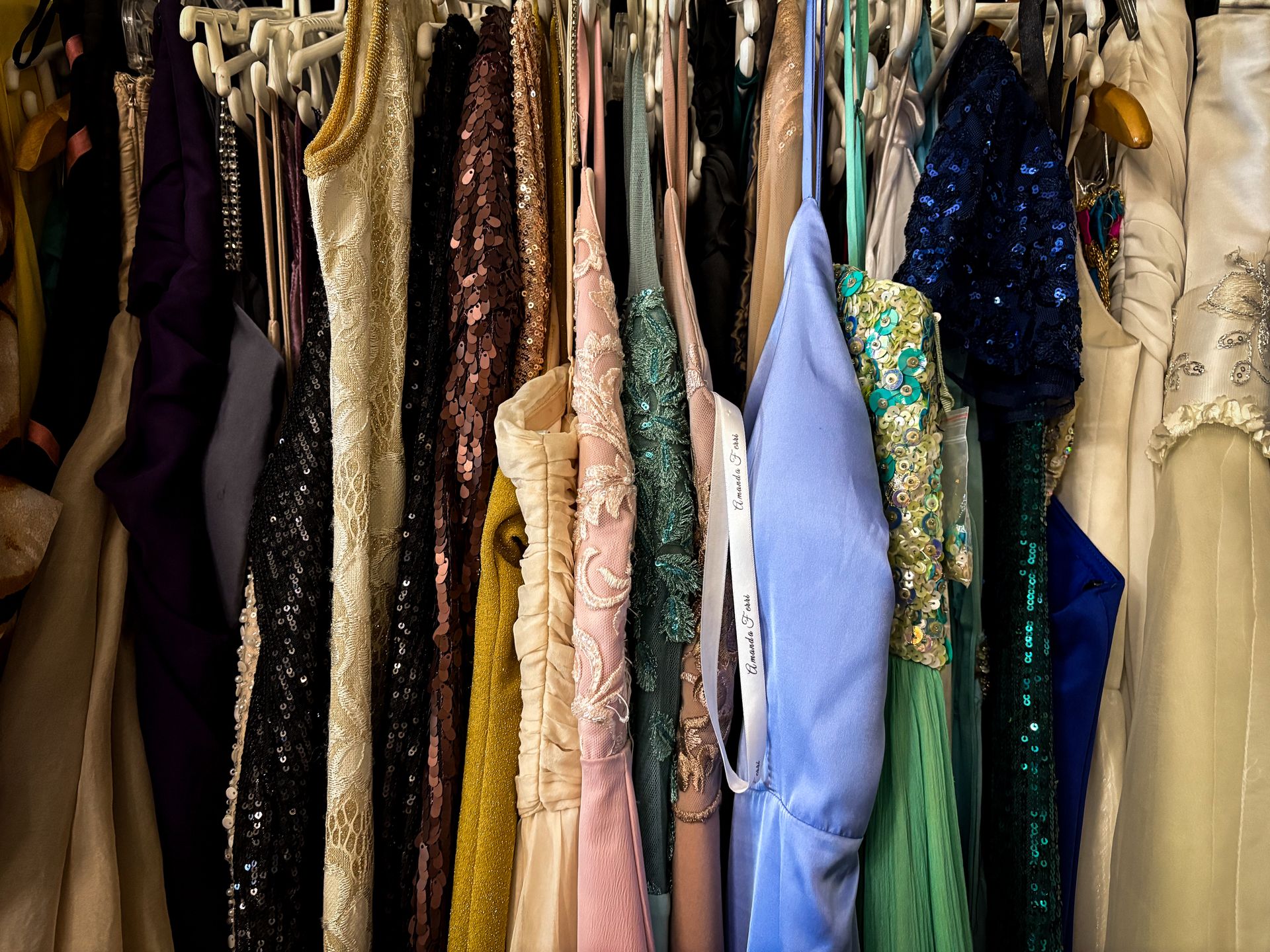 A rack of various formal gowns hanging closely together. The dresses feature diverse textures including sequins, lace, and solid satin in colors ranging from pastel pink and blue to black and gold.