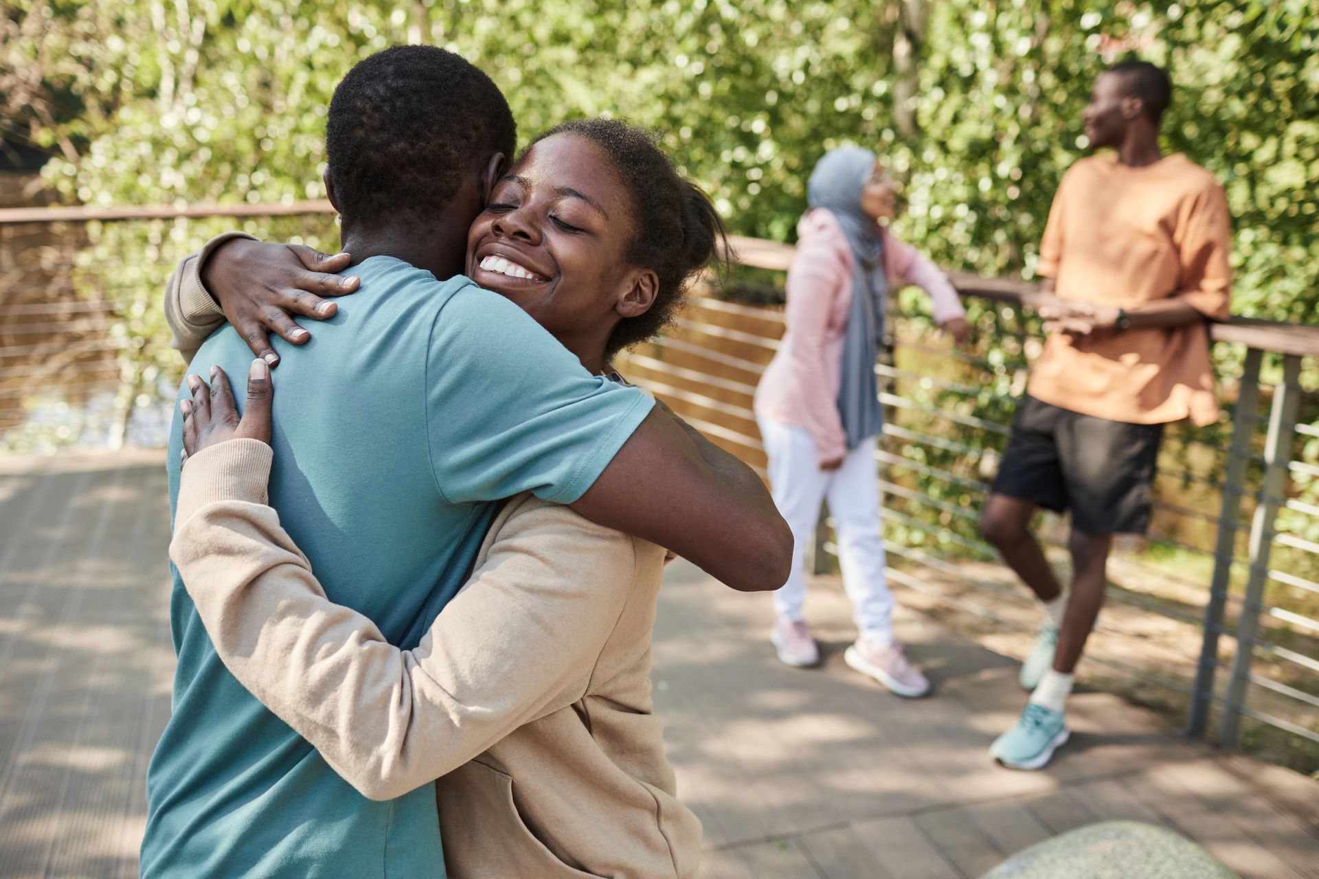 A young woman with her eyes closed smiles warmly while hugging a man on a wooden walkway. In the background, a man and a woman stand by a railing, talking.